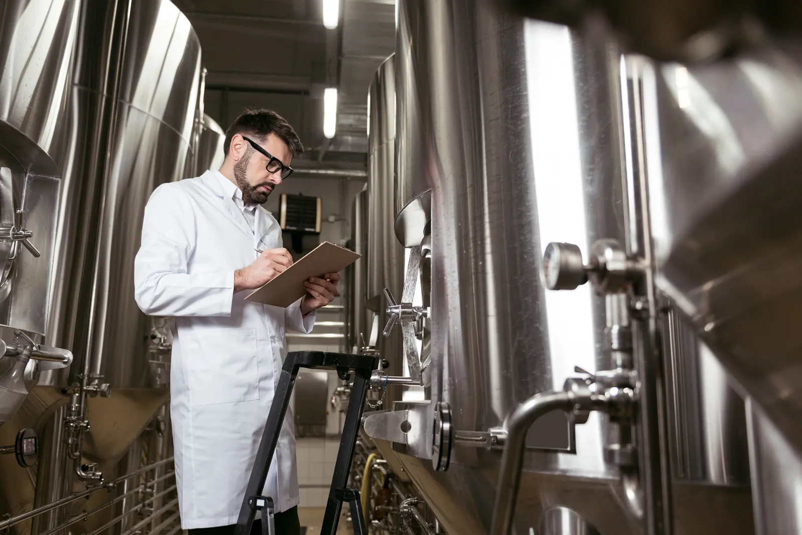 Engineer documenting measurements beside stainless steel processing vessels and sanitary process equipment.