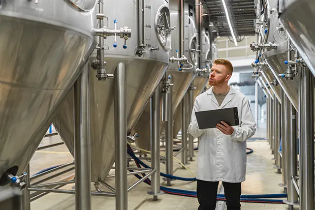 Quality control technician inspecting stainless steel mixing and processing tanks in a food and beverage production facility.
